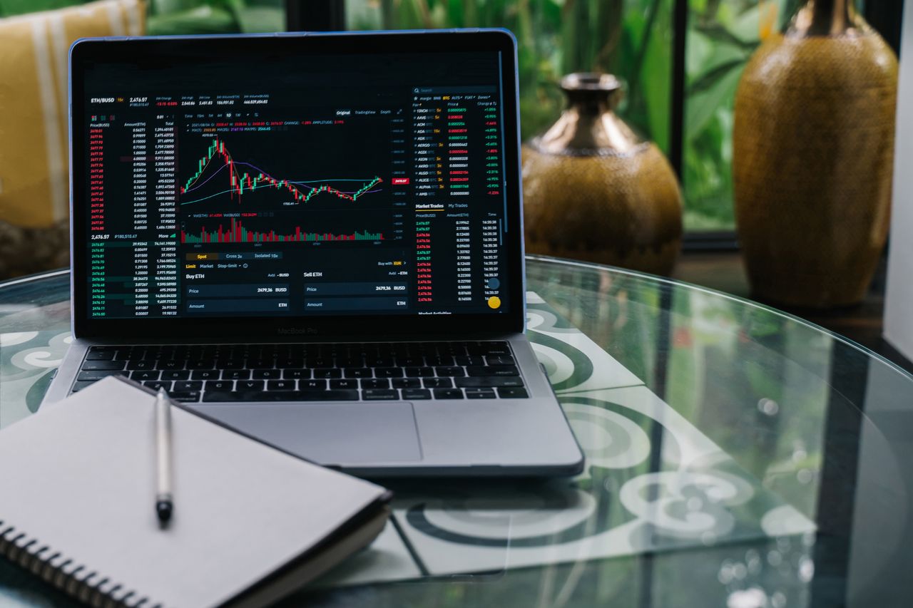 hero-img-01 Laptop displaying cryptocurrency stocks and graphs on a glass table with a notepad.